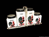 Front view of four milk glass spice shakers labeled Pepper, Flour, Sugar, Salt, each with a colorful rooster illustration, sitting in a worn metal caddy with handle.