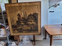 Folding occasional card table upright displaying sepia village scene artwork framed in a wooden border with some wear and minor damage.