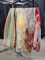 Five assorted light scarves hanging on black hanger arranged side by side showing diverse prints and colors including stripes, floral, animal print and geometric patterns.