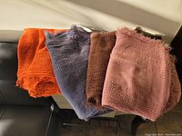 Four folded scarves in fall orange, slate blue grey, chocolate brown, and light pink colors arranged side by side on a black fabric surface.