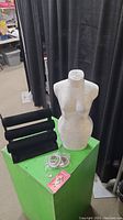 Silver style torso bust with textured surface alongside black velvet padded jewelry display rack, breast cancer keychain, and plastic toy bowls on a green pedestal against black curtain background.