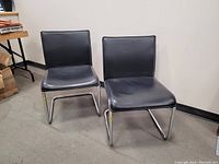 Pair of black upholstered mid century modern chairs with chrome cantilever frames placed against a plain wall on a dark floor.