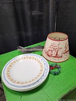 Photo of lampshade with sailing ship illustration, beaded bracelet in front, and metal cutlery behind stacked on green surface