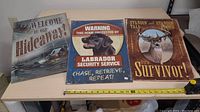 Three metal signs on a shelf showing a Labrador dog security sign, deer hunting sign, and cabin welcome sign