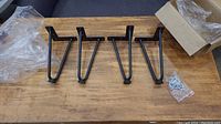 Four black metal hairpin legs with adjustable foot caps laid out on a wooden surface, showing shape and included hardware.