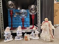 Three tall decorative goblets with red glass stems and bases, decorated with holly berry and leaf motifs on clear bowls visible on shelf with figurines