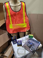 Full view of the lot items including safety vest on mannequin, measuring tapes, shaker, glasses, ashtray, vintage book, and box on white table