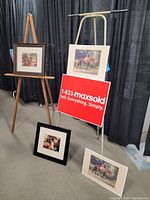 Wooden and metal easels supporting framed and unframed art prints including garden scenes and horse imagery, displayed against a black backdrop.
