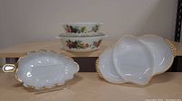 Photo of two English Pyrex JAJ milk glass baking dishes with vegetable pattern stacked behind two Fire-King white gold edge serving dishes on wooden surface, showing shape and pattern details.
