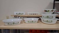 Display of 4 Glasbake milk glass bowls with green floral design and one square JAJ Pyrex brownie pan stacked and arranged on wooden shelves.