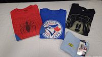 Four folded shirts lying on a white surface, showing red Spider-Man shirt, blue Toronto Blue Jays shirt, black Mickey Mouse shirt, and light blue NASCAR tank top with tag visible.