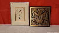 Photo shows two Indigenous art pieces side by side against a red backdrop: a framed print of a dancing loon with cracked glass (left) and a framed eagle artwork (right) approximately 12x12 inches.