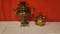 Both brass items side by side against orange background on white cloth. The larger samovar with ornate handles and spigot is on the left; the smaller covered pot with raised finial and three legs is on the right.
