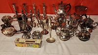 Wide view of assortment of vintage silverplate items showing various teapots, candlesticks, bowls, and serving items on a white surface with red background.