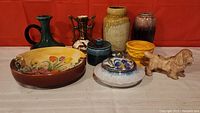 Photo showing the entire lot of 9 pottery items including vases, dish, lidded pot, and dog figurine arranged in front of red background.
