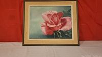 Framed painting of a pink rose with a light green background placed against a red backdrop.