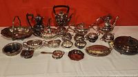 Full view of all silverplate items on table against a red backdrop, including various tea pots, serving trays, sugar bowl, creamers, and other pieces with tarnish visible.