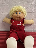 Front view of the vintage 1984 Cabbage Patch Kids doll seated with arms outstretched, showing blonde curly hair, blue eyes, facial freckles, and red corduroy overalls.