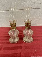 Two clear glass hurricane lamps with twisted bases and curved chimneys on a red surface against a beige wall.