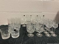 Full view of 15 glasses including tumblers, wine glasses and snifters all with pinwheel crystal patterns on black cloth.