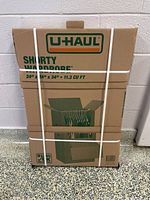 Photo of sealed cardboard box of U-Haul Shorty Wardrobe with green and orange logo and white strapping showing box dimensions and usage as a wardrobe box.