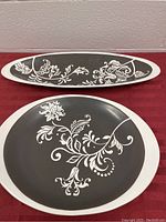 Two oval ceramic platters, one larger round style and one smaller long style with white floral and vine design on dark gray background.