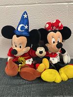 Three Disney plush toys: Sorcerer Mickey, classic Mickey, and Minnie Mouse, displayed together against a wall.