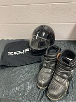 Black Zeus full-face motorcycle helmet with clear visor and drawstring pouch beside a pair of black Icon motorcycle boots size 11 with visible light wear.