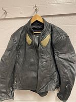 Front view of black perforated leather motorcycle jacket hanging on a hanger, showing shoulder padding and zippered cuffs.