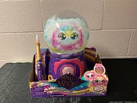 Full front view of Magic Mixie toy in packaging showing purple base, clear dome with colorful face inside, and wand on left.