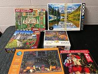 Photo showing six boxed jigsaw puzzles arranged on a grey surface against a white brick wall. Visible puzzles include Art Gallery, Rocky Mountains, Castle, artistic glitter themed, and still life with pumpkins.