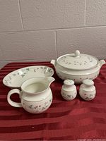 Tableware set on red cloth including covered serving bowl, gravy boat, salt and pepper shakers, and an oval serving dish. Floral strawberry design visible.