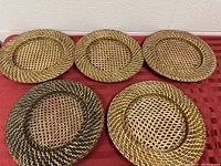 Five wicker rattan plate holders arranged on red cloth, showing natural and darker brown woven patterns for holding plates.