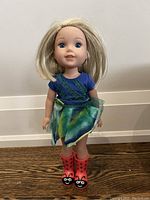 Front view of the Camille doll showing blonde hair, blue eyes, blue top, green sparkly skirt, and red lady bug rain boots.