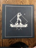 Black leather-like baseball card album binder with embossed baseball player and text, closed and laying flat on wood surface.