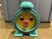 Front view of vintage Tommy The Time Out Timer Clock showing cartoon face with movable eyes, pink nose and teal plastic frame with numbered outer dial.