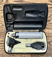 Top view of open case showing all three components: metal handle, otoscope head with black speculum, and ophthalmoscope head within molded insert.