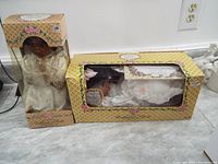 Two Crowne porcelain dolls in original boxes showing wear and damage; one doll dressed in cream lace, dark skin tone; other dressed in white satin with veil, light skin tone.