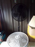 Black Lasko pedestal fan standing tall, missing one base support, against a metal wall background.