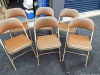 Group of six metal folding chairs with brown padded seats and backs outdoors, showing wear and folding design