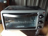 Front view of Black & Decker toaster oven on cardboard, showing stainless steel handle, glass door, three control dials on the right side, and inside removable tray and wire rack.