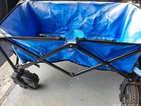 Side view of collapsible utility cart displaying the blue fabric interior and the black metal frame with large black tires, showing wear and scratches.