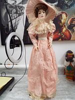 Full-length front view of the porcelain doll showing the pink satin gown with floral designs, lace collar, and wide-brimmed hat with tulle veil. Doll stands upright with hands at sides.