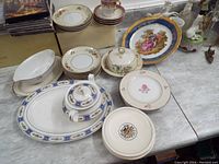 Overview photo showing entire ceramic lot including serving platters, plates, tea cup with saucer, lidded butter dish, and decorative plates on table.
