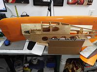 Model airplane parts including orange wing and bare wooden fuselage frame on a table with instructions.