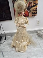 Full body view of porcelain doll standing on floor near wall outlet, dressed in cream satin vintage dress with puffed sleeves and large hat with veil