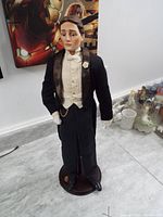 Full front view of porcelain groom doll standing on wooden base, dressed in tuxedo with white gloves and boutonniere.