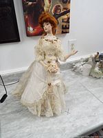 Full view of porcelain doll standing on floor with light beige Victorian lace dress and red curly hair.