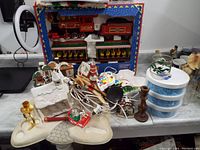 Various assorted Christmas decorations including wood train ornaments, Santa figurines, candle holders, glassware, and storage containers, displayed on a table