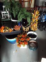 Wide view of multiple artificial plants, planters, and pots showing variety of sizes, colors and materials.